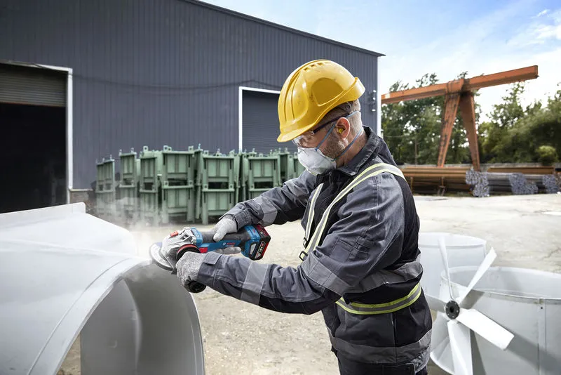 Une personne portant un équipement de sécurité broie du métal à l’extérieur sur un site industriel.