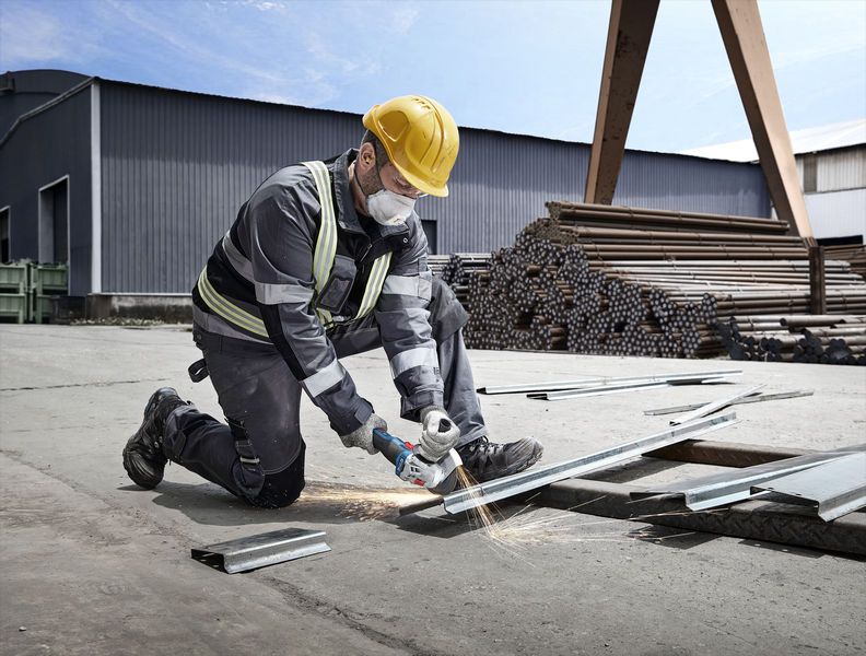 Un travailleur portant un équipement de sécurité coupe du métal avec une meuleuse électrique sur un site industriel.