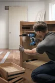 Une personne portant un équipement de sécurité utilise une perceuse sans fil pour assembler une armoire en bois.