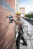 Une personne portant un équipement de sécurité perce un mur de briques tout en se tenant debout sur un échafaudage.