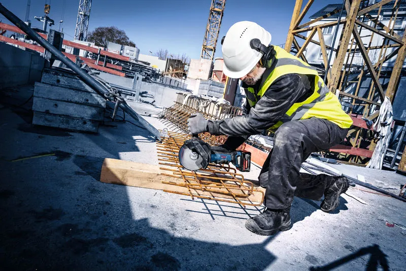 Une personne portant un équipement de sécurité coupe des barres d'armature sur un chantier de construction avec une meuleuse d'angle sans fil.