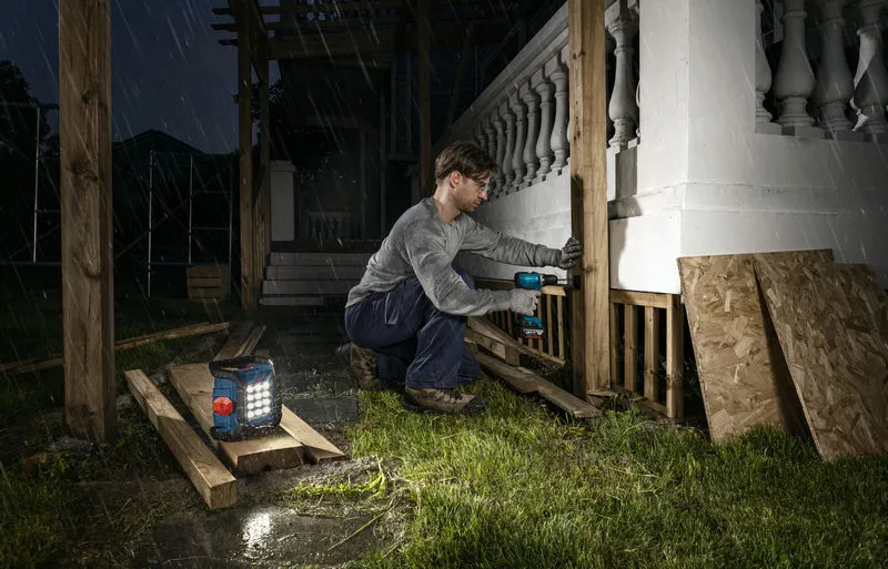 Une personne portant un équipement de sécurité perce des poteaux en bois la nuit avec une lampe de chantier sans fil à proximité.