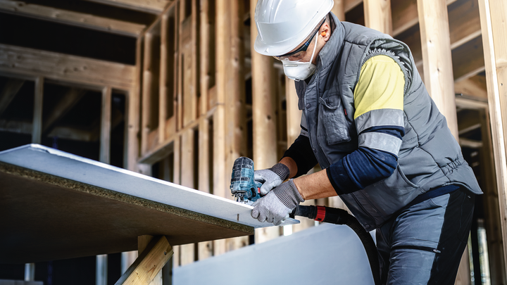 Une personne portant un équipement de sécurité utilise une perceuse électrique sur une plaque de plâtre dans une ossature de bâtiment.
