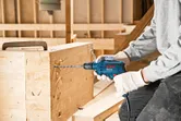 Une personne portant un équipement de sécurité perce une poutre en bois à l'aide d'une perceuse électrique.