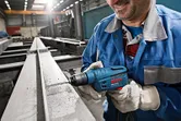 Une personne portant un équipement de sécurité perce une poutre en acier à l'aide d'un outil électrique bleu.
