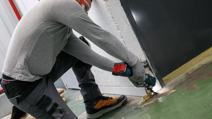 Une personne portant un équipement de sécurité utilise un outil électrique sans fil pour retirer le revêtement de sol près d'un mur.