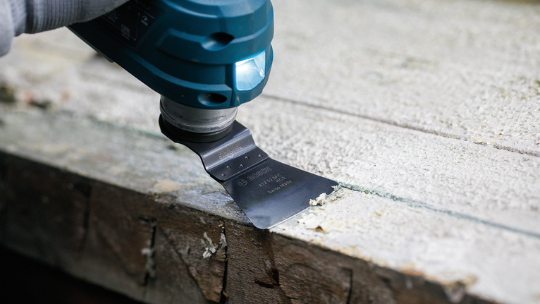 Une personne portant un équipement de sécurité utilise un outil multifonction pour gratter des matériaux du bois.