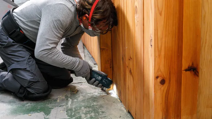Une personne portant un équipement de sécurité utilise un outil électrique pour couper près d'un mur en bois.