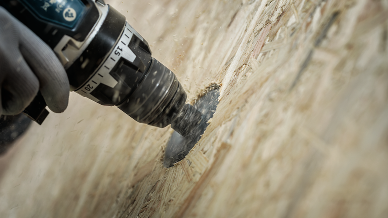 Une personne portant un équipement de sécurité perce un trou circulaire dans du contreplaqué.