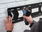 Une personne fixe un support métallique sur un mur de briques blanches à l'aide d'une perceuse sans fil.