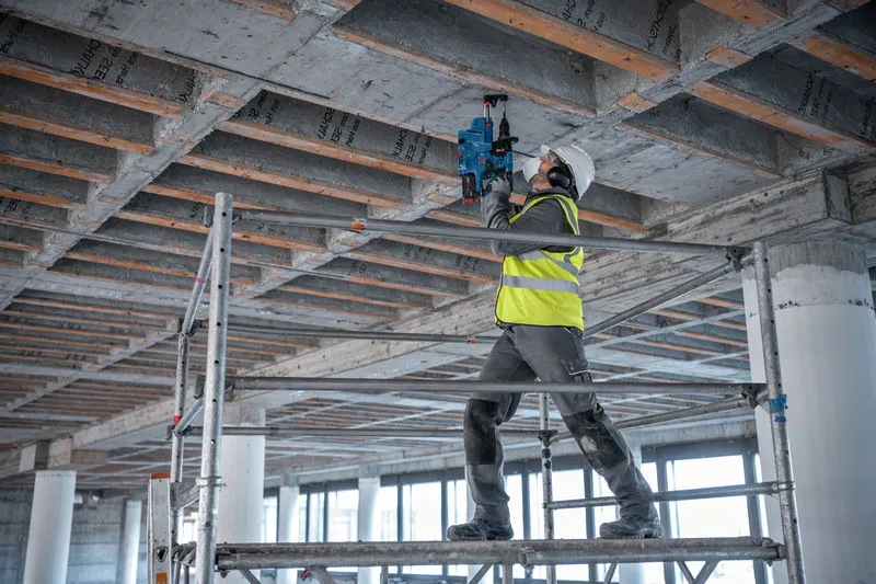 Un ouvrier portant un équipement de sécurité utilise un marteau perforateur pour percer un plafond en béton.