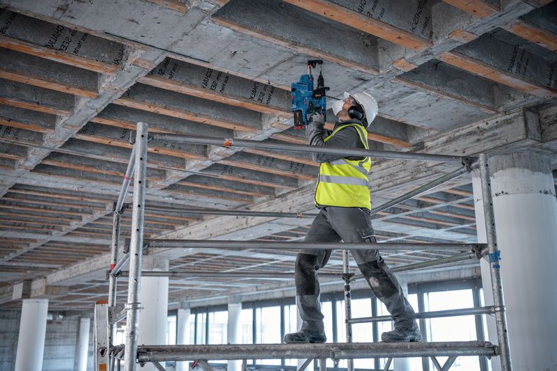 Un ouvrier portant un équipement de sécurité utilise un marteau perforateur pour percer un plafond en béton.