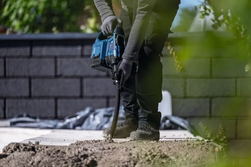 Une personne portant un équipement de sécurité utilise un outil électrique pour percer du béton à l'extérieur.