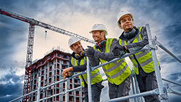 Drie arbeiders in gele vesten en helmen, voor een gebouw in aanbouw, glimlachend.