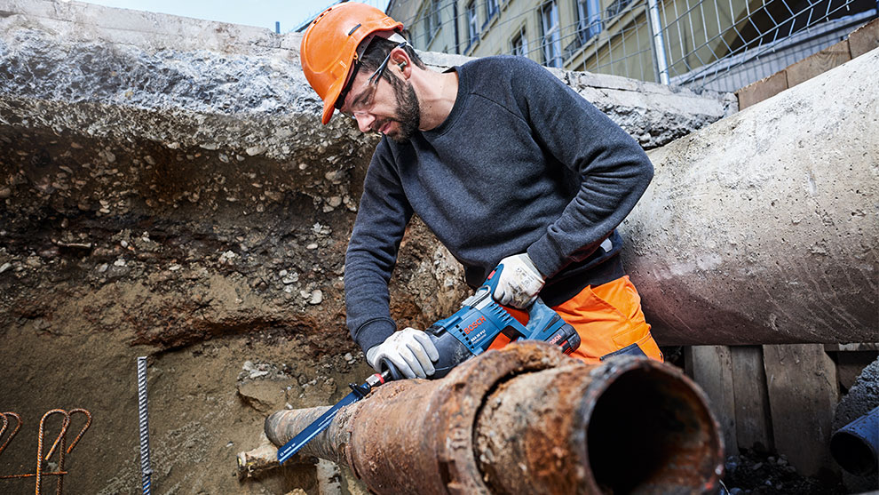 Een man met een oranje helm gebruikt een Bosch-boormachine op een verroeste pijp.