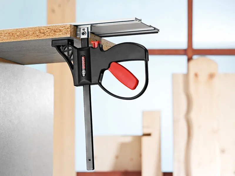 Systeemklem waarmee een houten plank aan een metalen oppervlak in een werkplaats wordt bevestigd.