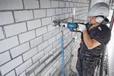 Een persoon met veiligheidsuitrusting boort met een diamantboor in een bakstenen muur.