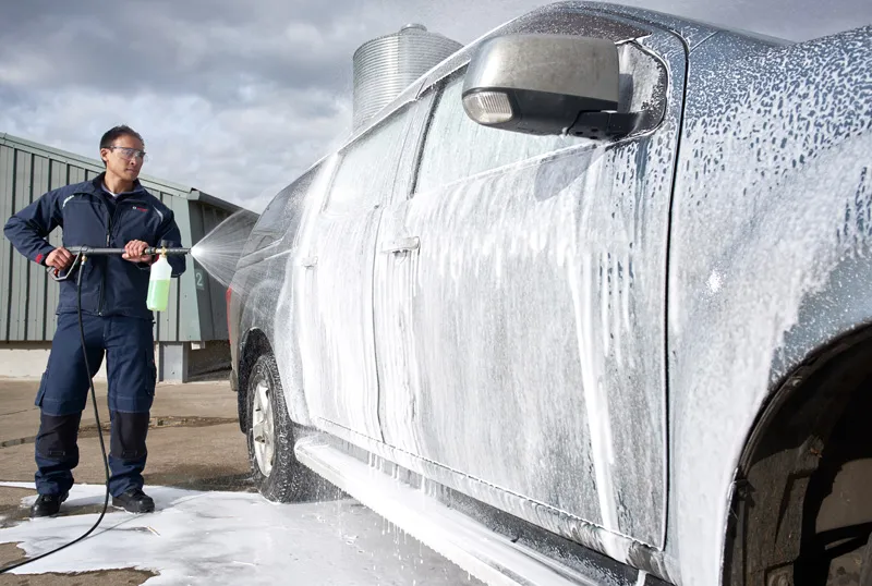Een persoon die veiligheidsuitrusting draagt, gebruikt een hogedrukreiniger om de buitenkant van de schuimende auto schoon te maken.