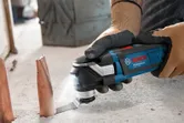 Een persoon die veiligheidsmateriaal draagt, gebruikt een multi-cutter om een metalen pijp op een betonnen vloer door te snijden.