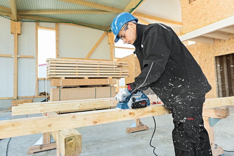 Een persoon die veiligheidsuitrusting draagt, gebruikt een schaafmachine om een houten balk glad te maken.
