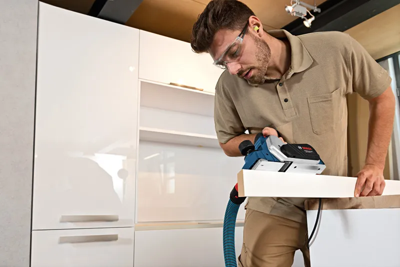 Een persoon die veiligheidsuitrusting draagt, gebruikt een schaafmachine op een houten plank.