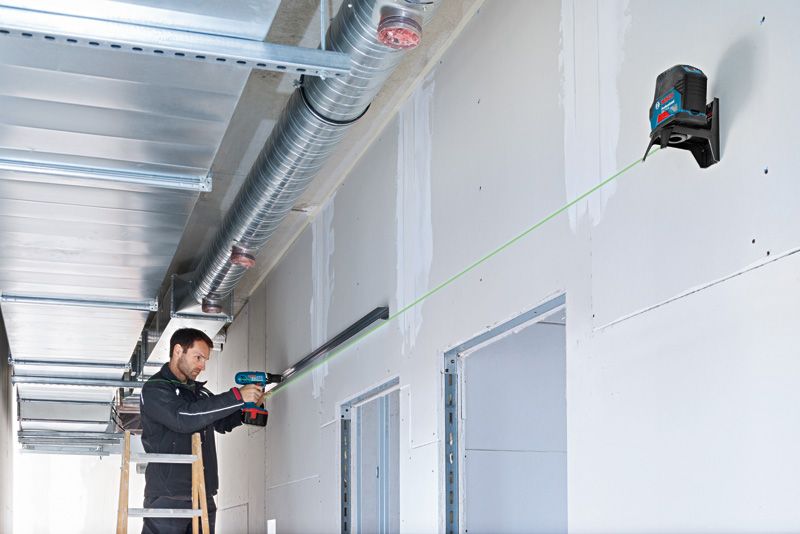 Een persoon monteert met een boormachine een metalen rail, geleid door een groene laserwaterpas.