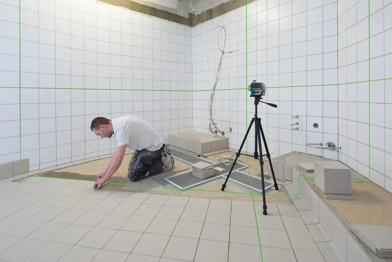 Persoon die tegels in een badkamer installeert, gebruikt een laserwaterpas op een statief.