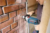 Een persoon met veiligheidsuitrusting boort met een slagboormachine in een bakstenen muur.