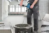 Een persoon die veiligheidsmateriaal draagt, mengt cement in een emmer met behulp van een elektrische mixer.