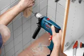 Iemand boort een schroef in een houten blok dat op een betegelde muur is bevestigd.