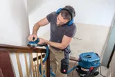 Een persoon met veiligheidsuitrusting schuurt een houten leuning met een excentrische schuurmachine.
