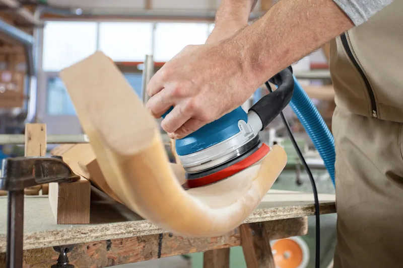 Persoon schuurt een gebogen onderdeel van een houten stoel met een excentrische schuurmachine.