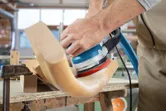 Persoon schuurt een gebogen onderdeel van een houten stoel met een excentrische schuurmachine.