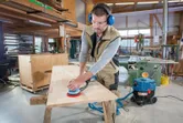 Een persoon met veiligheidsuitrusting schuurt een houten tafelblad met een excentrische schuurmachine.