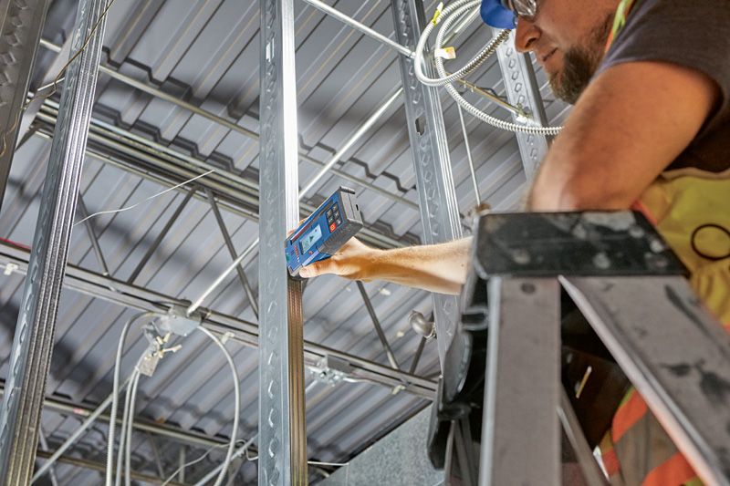 Een persoon die veiligheidsuitrusting draagt, gebruikt een laserontvanger op een metalen balk in een gebouw.