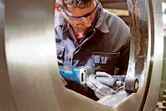 Een persoon met veiligheidsuitrusting polijst een grote metalen ring met een snoerloze rechte slijper.
