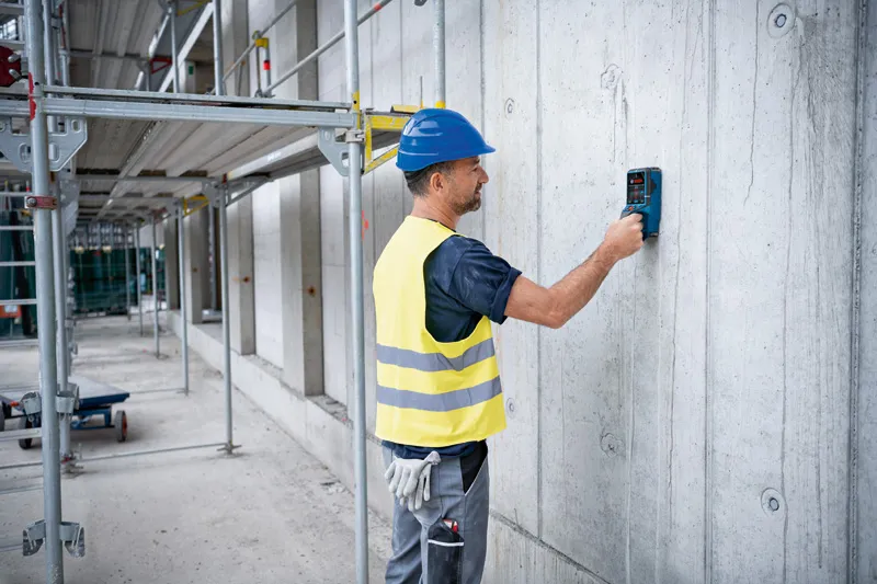 Een persoon met veiligheidsuitrusting scant een betonnen muur met een detectorinstrument.