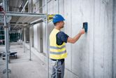 Een persoon met veiligheidsuitrusting scant een betonnen muur met een detectorinstrument.