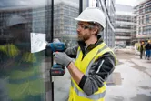 Een persoon die veiligheidsuitrusting draagt, gebruikt een accuboormachine op een glazen gevel.