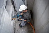Een persoon met veiligheidsuitrusting boort in een muur om een ​​oranje buis te installeren.