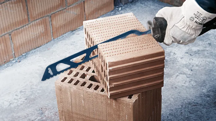 Een persoon met veiligheidsuitrusting zaagt een bakstenen blok door met een handzaag.