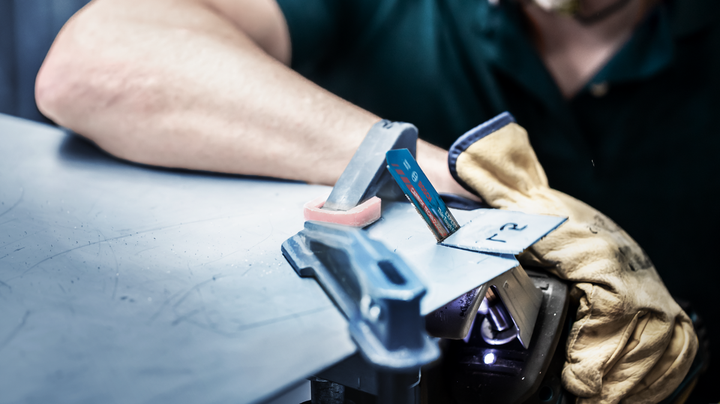 Een persoon die veiligheidsuitrusting draagt, houdt een metalen plaat vast met een handschoen terwijl hij een decoupeerzaag gebruikt.