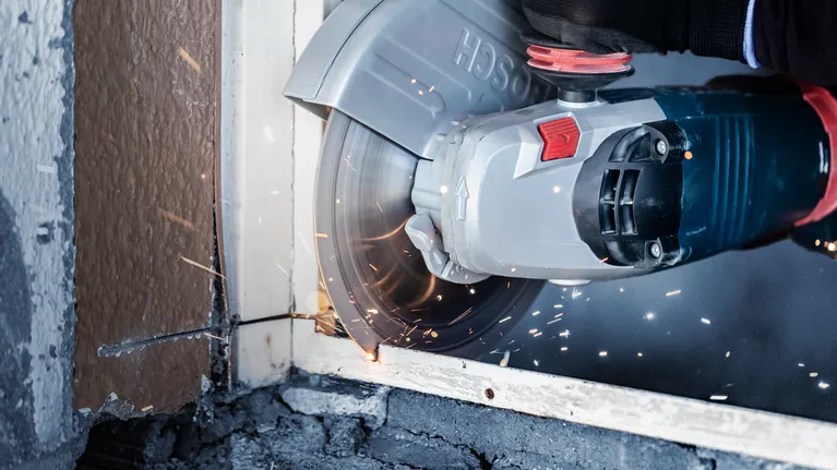 Persoon met veiligheidsuitrusting zaagt een metalen raamkozijn door met een grote haakse slijper.