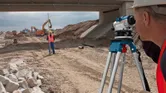 Een persoon die veiligheidsuitrusting draagt, gebruikt een optische waterpas op een wegwerkzaamhedenterrein.