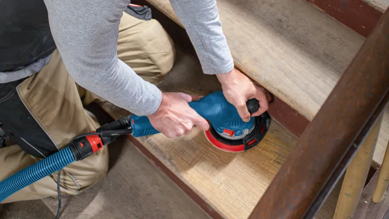 Een persoon schuurt houten trappen met een excentrische schuurmachine die is aangesloten op een stofzuigerslang.