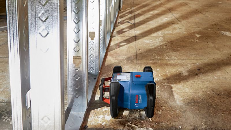 Laserwaterpasinstrument projecteert richtlijnen naast metalen wandframes op een bouwvloer.