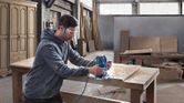 Een persoon die veiligheidsuitrusting draagt, gebruikt een bovenfrees op een houten tafelblad in een werkplaats.