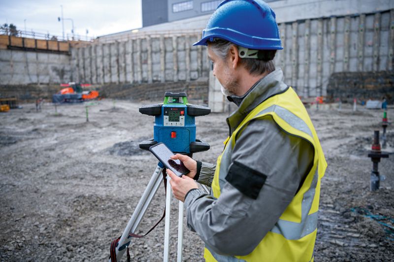 Een persoon die veiligheidsuitrusting draagt, bedient een laserwaterpas op een bouwplaats.