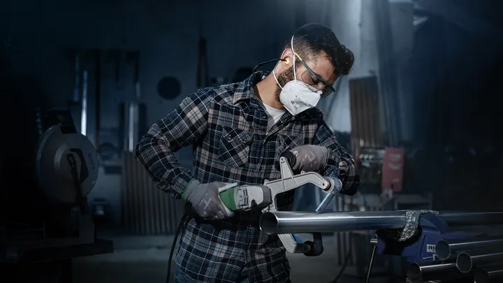 Een persoon met veiligheidsuitrusting slijpt een metalen pijp in een werkplaats.