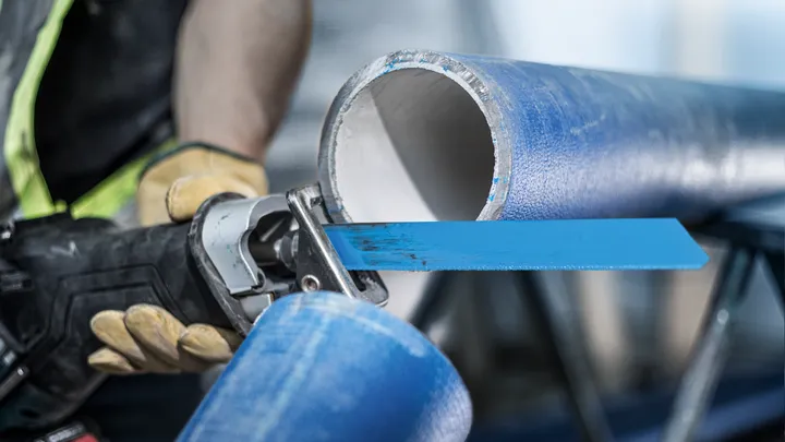Een persoon met veiligheidsuitrusting zaagt een grote blauwe pijp door met een reciprozaag.
